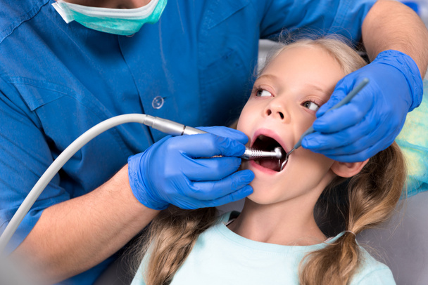 abgeschnittene Aufnahme eines Zahnarztes mit einem Zahnhandstück, das mit Zähnen eines kleinen Kindes arbeitet - Foto, Bild