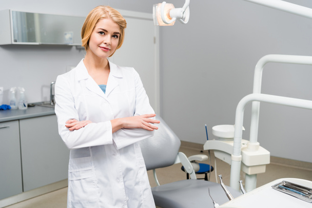 atractivo joven dentista con brazos cruzados mirando a la cámara en la oficina
 - Foto, Imagen