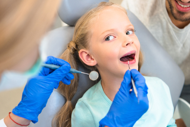bijgesneden schot van vrouwelijke tandarts behandeling van tanden van klein kind terwijl vader zitten in de buurt van haar - Foto, afbeelding