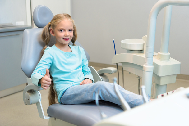 sorridente bambino seduto sulla sedia dentale presso lo studio dentistico e mostrando pollice in su
 - Foto, immagini