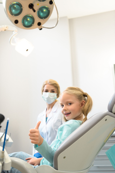 niño sonriente mostrando el pulgar hacia arriba mientras está sentado en la silla en el consultorio del dentista
 - Foto, Imagen