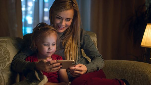 Mother and Her Cute Daughter are Sitting On a Couch in the Living Room. They're Using Smartphone and Smiling. It's Evening. - Фото, изображение