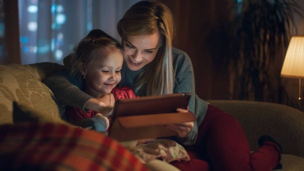 Beautiful Mother and Her Little Daughter are Sitting on a Sofa in the Living Room, They Use Tablet Computer. It's Evening, Room is Cozy and Warm. - Фото, изображение