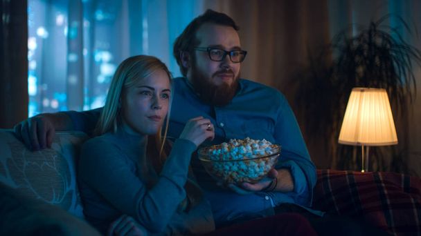 Boyfriend and Girlfriend Watching TV. They Sit on a Sofa in Their Cozy Living Room and Eat Popcorn. It's Evening. - Zdjęcie, obraz