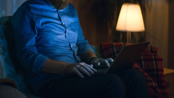 Close up shot of Man Works on His Laptop While Sitting on Sofa in His Living Room. É noite.
. - Foto, Imagem