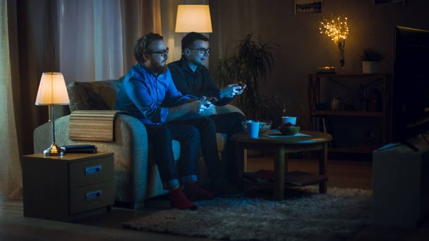 In the Evening Two Friends are Sitting on a Sofa in the Living Room and  Playing Competitive Video Games. They Push and Shove Each Other Friendly. - Foto, imagen