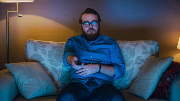 Portrait Shot of a Man Sitting on a Sofa in His Living Room, Eating Popcorn and Watching TV. Floor Lamps are Turned ON. - Foto, imagen