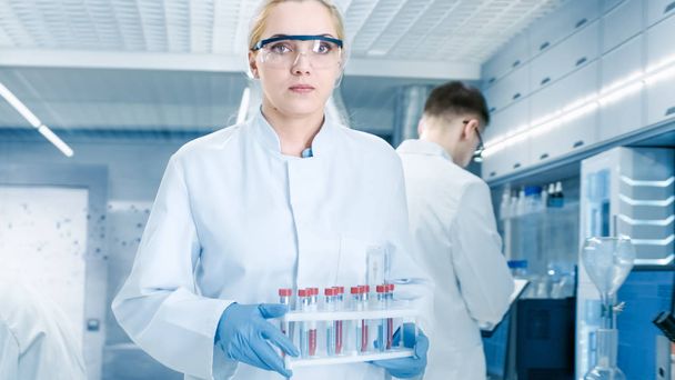 Shot of Young Female Scientist Walking with Case of Test Tubes Through Busy Laboratory. Male and Female Scientists of Different Age Working with Technical Equipment. - Photo, Image