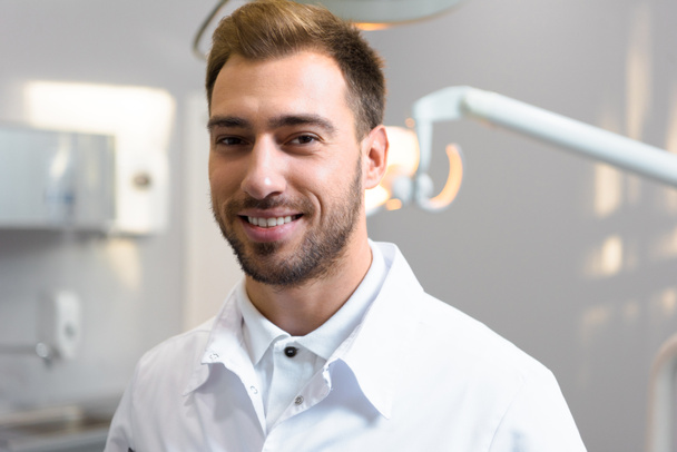 primer plano de sonriente joven dentista en abrigo blanco mirando a la cámara en la oficina
 - Foto, Imagen