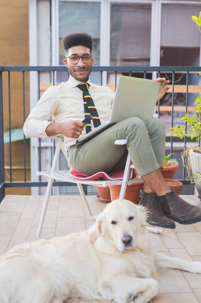 młody czarny człowiek kryty siedzi kanapa za pomocą laptopa i smartfona - zdalna praca, biznes, koncepcja technologii - Zdjęcie, obraz