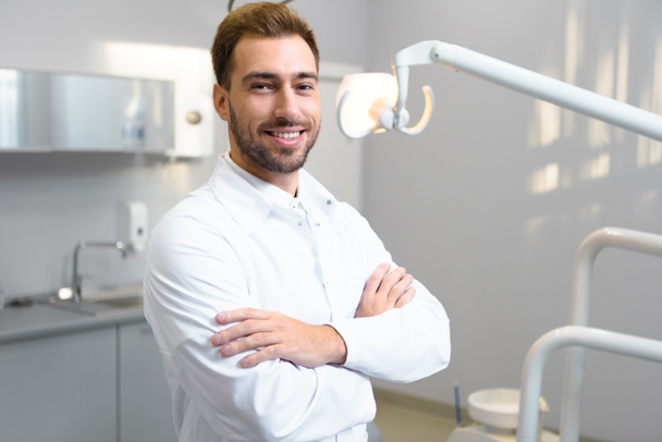 przystojny młody stomatolog ze skrzyżowanymi rękami patrząc na kamery w biurze - Zdjęcie, obraz