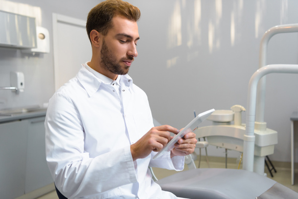 handsome young dentist in coat using tablet in office - Zdjęcie, obraz