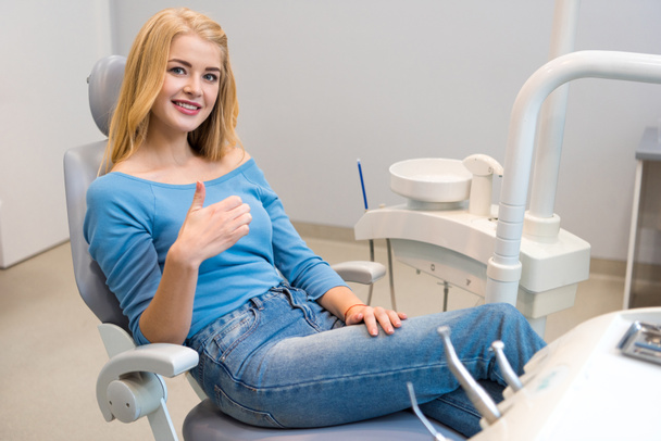 boldog, fiatal nő, a fogorvosi rendelőben a fogorvosi székben ül, és felbukkan a hüvelykujj - Fotó, kép