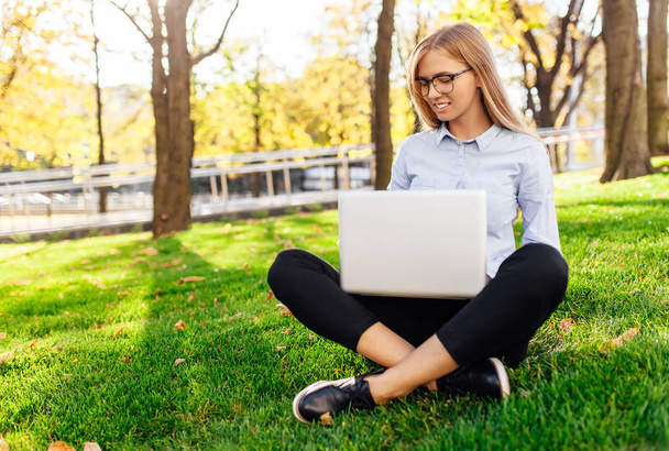 Young attractive girl with glasses, Woman sitting on the grass, working on a laptop, in a city park on a green lawn outdoors. Freelance business concept - 写真・画像