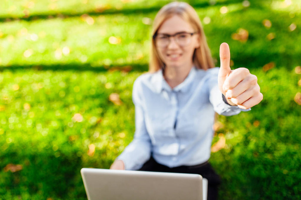 Bild einer jungen Geschäftsfrau, die in einem Park sitzt, einen Laptop benutzt und eine Geste zeigt, Klasse, auf einem grünen Rasen sitzend. - Foto, Bild