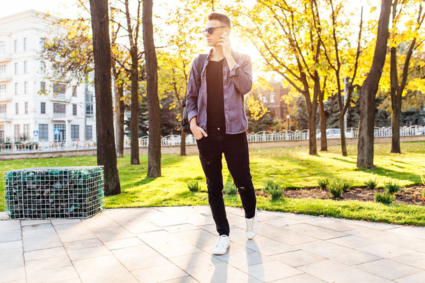 young guy dressed in stylish clothes, listening to music through wireless headphones and walking in the park, outdoors - Foto, Imagen