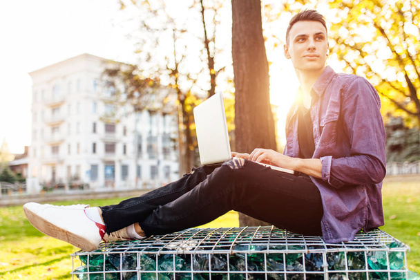 man, in casual clothes, sits on a bench, works with a laptop and listens to music using wireless headphones, against the backdrop of the sunset - Foto, imagen