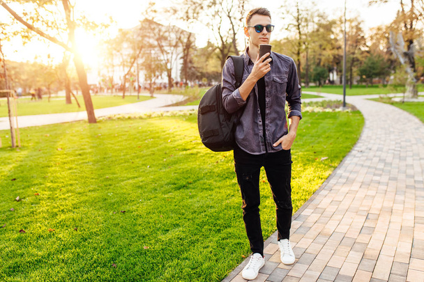 attractive man with a briefcase, dressed in stylish clothes, sunglasses, holds in his hand a glass of coffee and walks in the park, - Фото, зображення