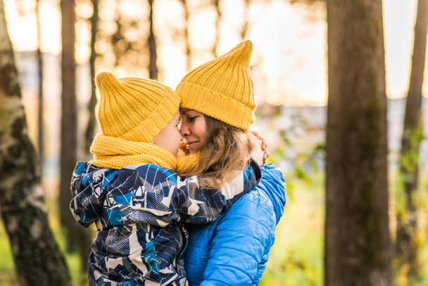 mom in a yellow hat and a blue jacket hugs her son in a yellow hat and a blue jacket autumn forest - Fotó, kép