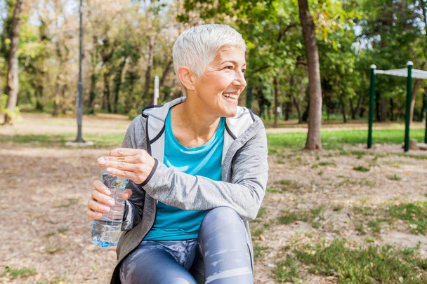 Mujer sana mayor Relájese y sostenga la botella de agua después del entrenamiento en el parque al aire libre de la aptitud
 - Foto, Imagen