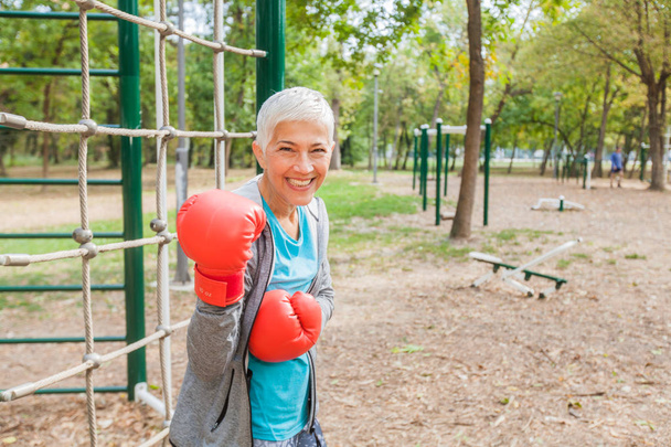 スポーツウェアの屋外フィットネス公園でボクシング グローブを合わせる年配の女性の肖像画。アクティブな昔の人は健康的なライフ スタイル - 写真・画像