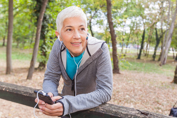 Zdrowa kobieta Senior zrelaksować się słuchając muzyki z telefonu po joggingu w lesie. Styl życia ludzi dojrzałych ćwiczenie odkryty. - Zdjęcie, obraz