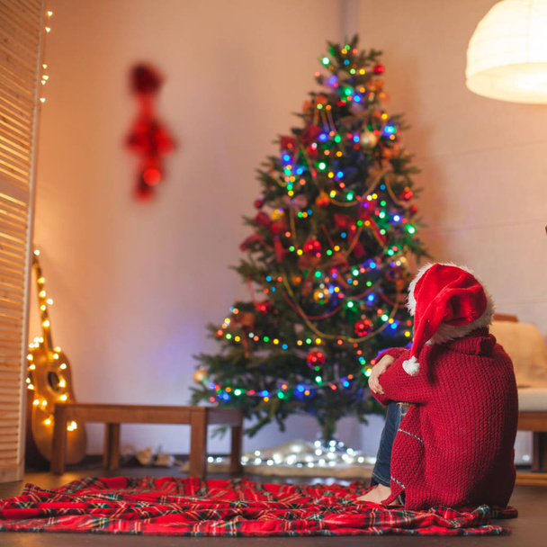 Το κορίτσι κάθεται με την πλάτη της και κοιτάζει το χριστουγεννιάτικο δέντρο - Φωτογραφία, εικόνα