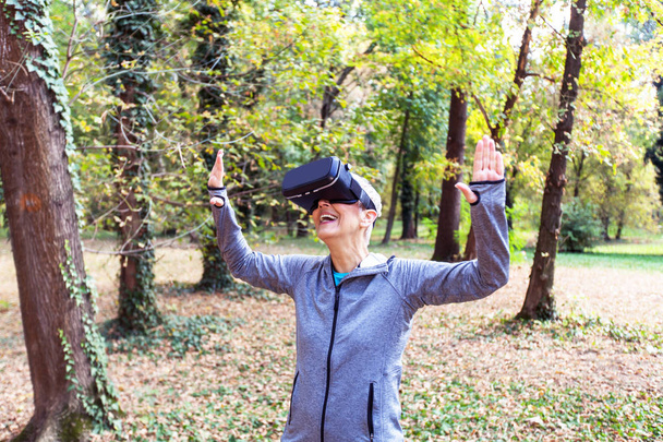 Счастливая пожилая женщина развлекается гарнитурой виртуальной реальности в лесу, прибором для пожилых людей
. - Фото, изображение