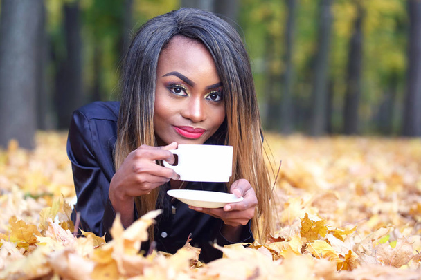 Piękna młoda Afrykańska kobieta picie kawy z białego Pucharu na tle jesiennych liści w par - Zdjęcie, obraz