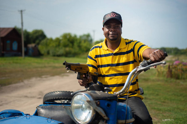 RUSSIA, SYZRAN - SEPTEMBER 2, 2018: black gangster with a gun in his hands on a motorcycle in the countryside - Foto, afbeelding