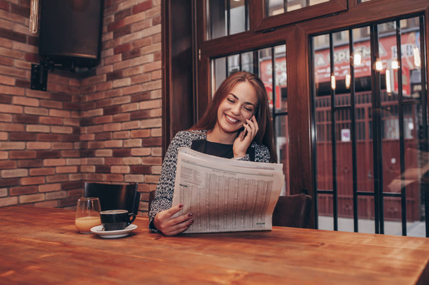Νέοι άνεργοι θηλυκό κάνει δουλειά κυνήγι και διαβάζοντας ανακοινώσεις απασχόλησης. Η γυναίκα κάθεται στο καφενείο κρατώντας μια εφημερίδα και να κάνει ένα τηλεφώνημα για να οργανώσει μια συνέντευξη. - Φωτογραφία, εικόνα