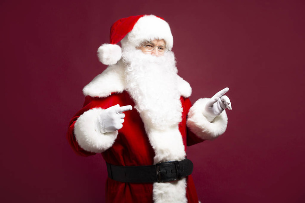 portrait of man in Santa Clause costume pointing by fingers aside while posing on red background, Christmas and New year concept  - Foto, imagen