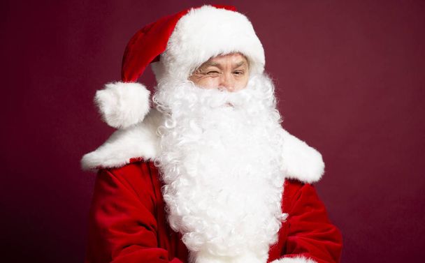  funny joyful man in Santa Clause costume  looking at camera and winking on red background, Christmas and New year concept  - Photo, Image