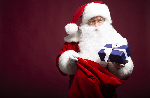 Mann im Weihnachtsmann-Kostüm schenkt Geschenk und hält mit Geschenksack auf rotem Hintergrund, Weihnachts- und Neujahrskonzept  - Foto, Bild