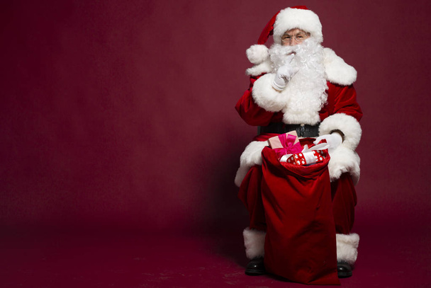 portrait of man in Santa Clause costume sitting with gift sack and showing silence sign on red background, Christmas and New year concept  - Valokuva, kuva