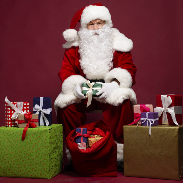 Mann im Weihnachtsmann-Kostüm beschenkt im Sitzen mit Geschenksack auf rotem Hintergrund, Weihnachts- und Neujahrskonzept  - Foto, Bild