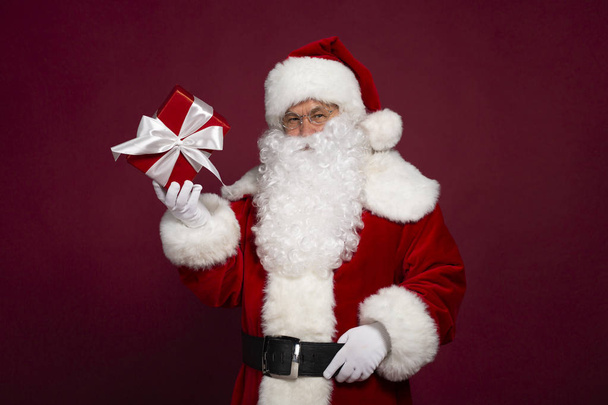 portrait of man in Santa Clause costume holding paper gift box and looking at camera on red background, Christmas and New year concept  - Fotoğraf, Görsel