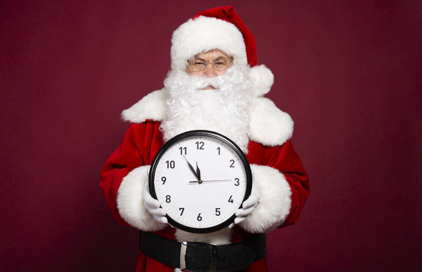portrait of man in Santa Clause costume holding white clock on red background, Christmas and New year concept  - Foto, imagen