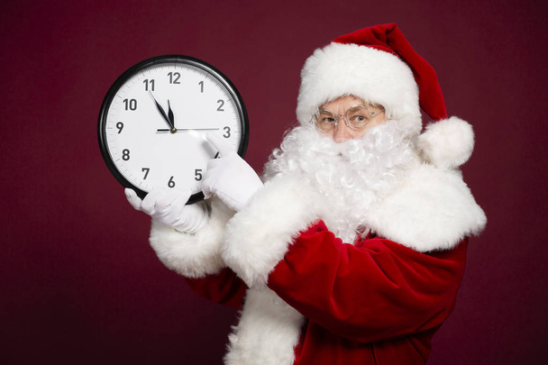 portrait of man in Santa Clause costume pointing at white clock in hand while posing on red background, Christmas and New year concept  - Foto, imagen