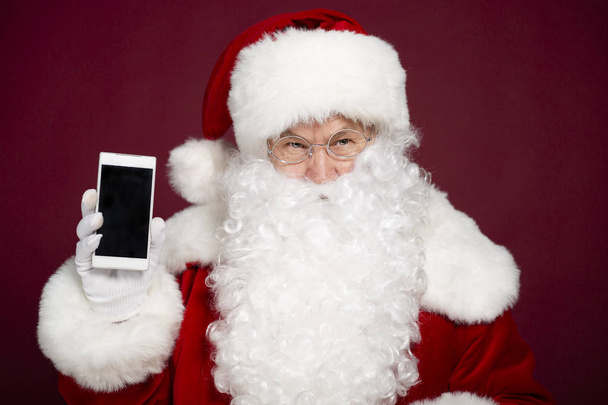 man in Santa Clause costume holding smartphone on red background, Christmas and New year concept  - Фото, изображение