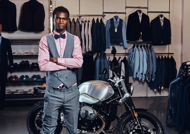 Elegantemente vestido joven afroamericano posando con brazos cruzados cerca de moto deportiva retro en la tienda de ropa para hombres
. - Foto, imagen