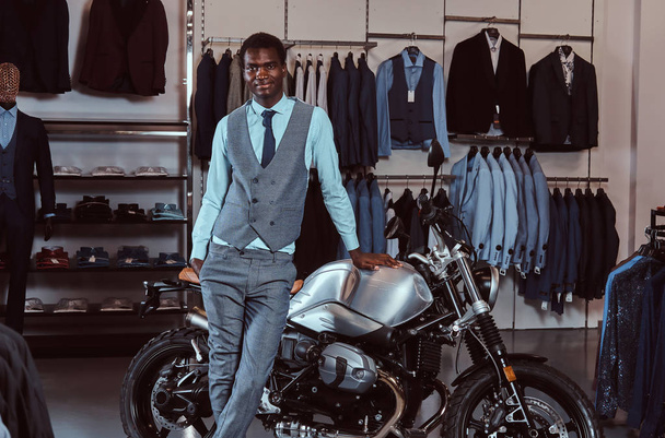 Elegancko ubrany African American młody mężczyzna pozowanie w pobliżu retro sportowy motocykl sklep Odzież męska. - Zdjęcie, obraz