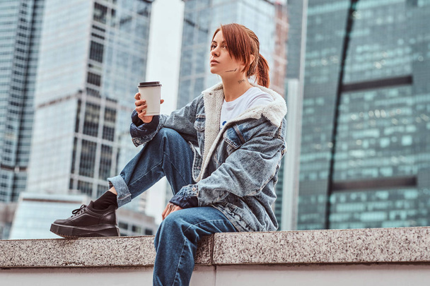 stylisches rothaariges Hipstermädchen mit Tätowierung im Gesicht, das Jeansmantel trägt und Kaffee zum Mitnehmen vor Wolkenkratzern in der Moskauer Stadt am trüben Morgen hält. - Foto, Bild