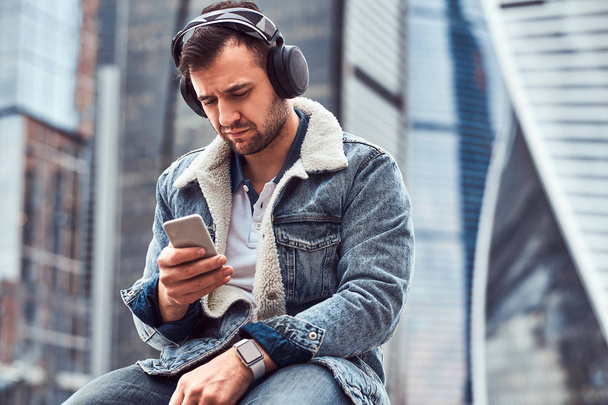 Stijlvolle man met denim jasje luisteren naar muziek en het gebruik van de smartphone zit van wolkenkrabbers in Moskow stad op bewolkte ochtend. - Foto, afbeelding