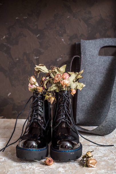 Yellow and pink flowers grow in the old black boots for original decorate. Lacquer black shoes with high soles. Gray felt bag. Concept of warm autumn or winter. copy space - 写真・画像