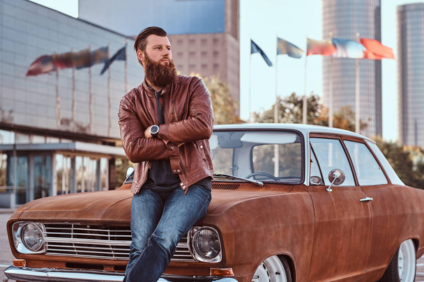 Bearded male dressed in brown leather jacket and boots leaning on tuned retro car in the city parking near skyscraper. - Foto, Imagen