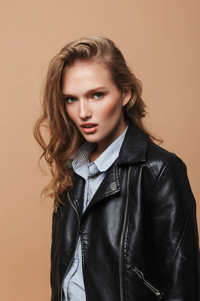 Young beautiful woman with wavy hair in black leather jacket and shirt thoughtfully looking in camera over beige background - Photo, Image