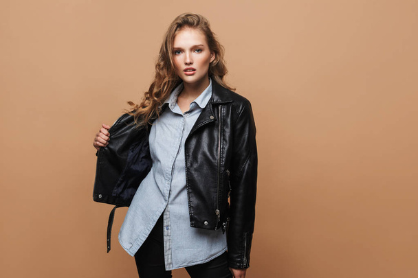 Young pretty woman with wavy hair in black leather jacket and shirt thoughtfully looking in camera over beige background - Photo, Image