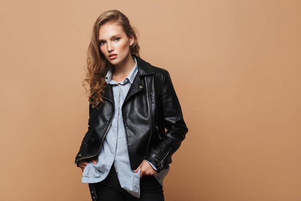 Young attractive serious woman with wavy hair in black leather jacket and shirt holding hands in pockets thoughtfully looking in camera over beige background - Photo, Image