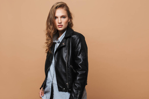 Young attractive woman with wavy hair in black leather jacket and shirt thoughtfully looking in camera over beige background - Photo, Image